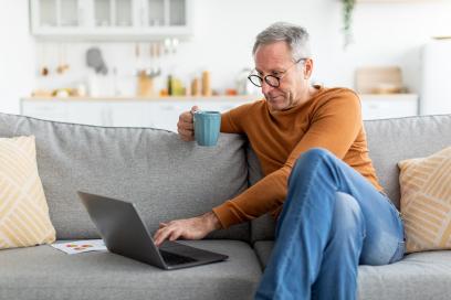 Älterer Mann sitzt mit Kaffee auf der Couch und ließt etwas am Laptop